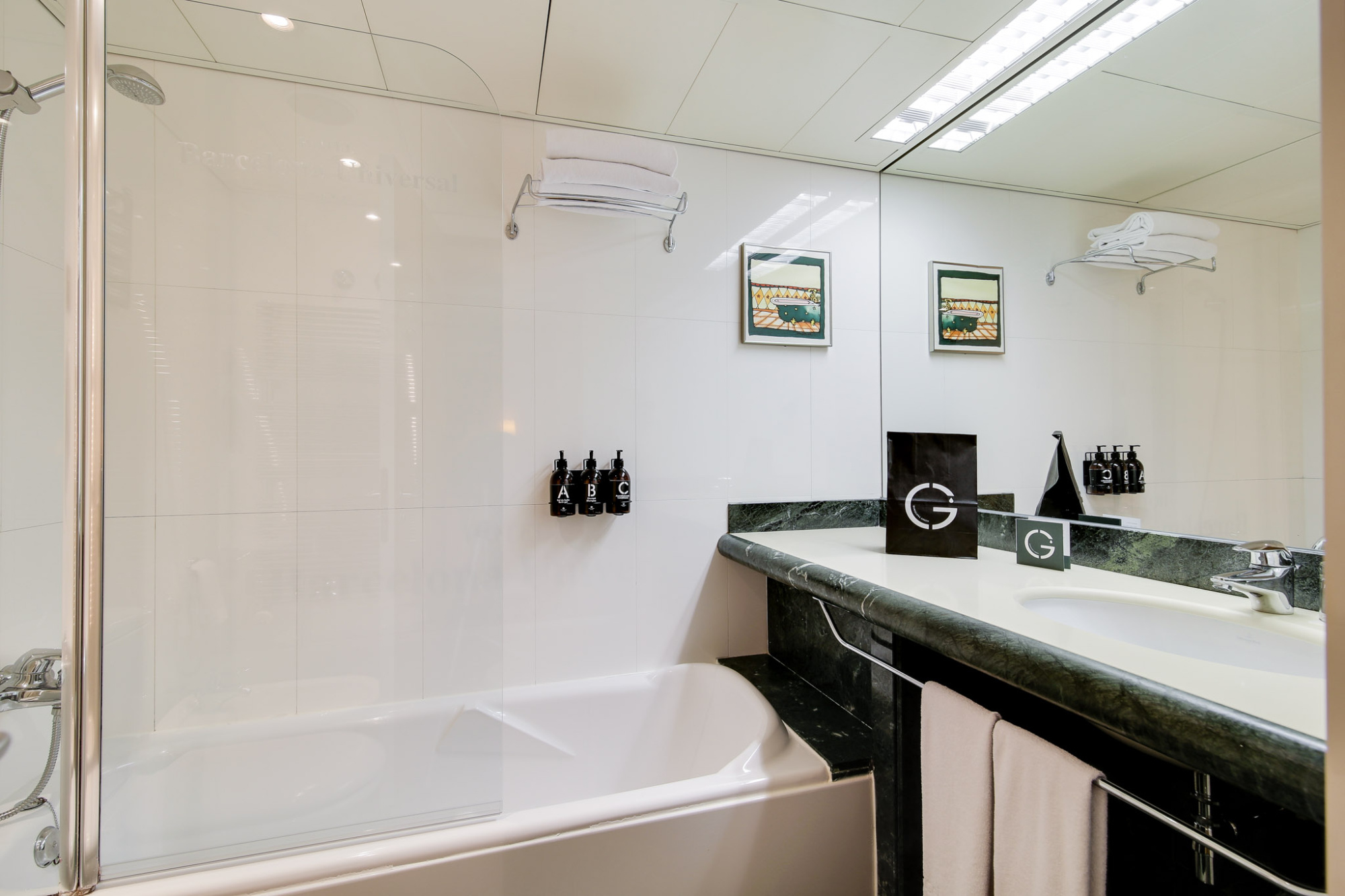 Se ve una bañera con mampara y un lavabo doble con toallas en un baño luminoso.