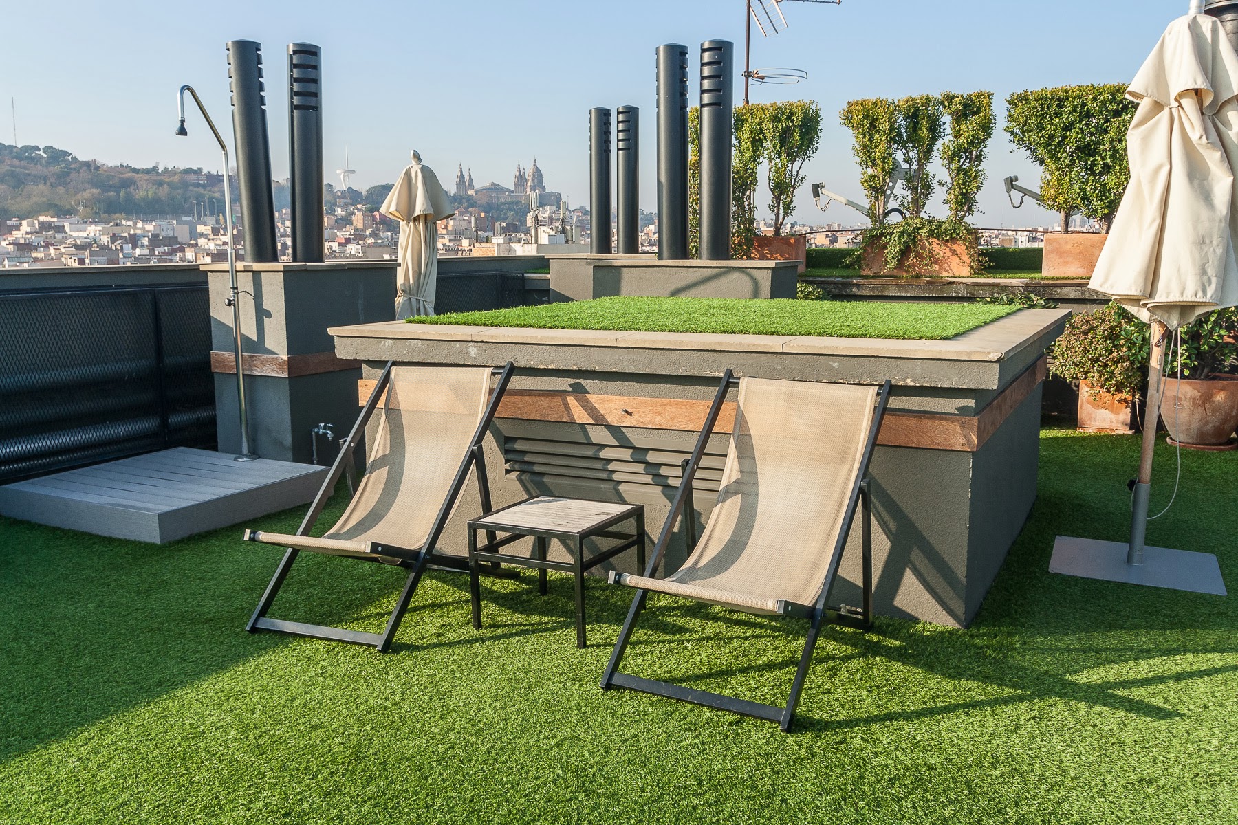 Se ve una terraza exterior con césped artificial, dos tumbonas y sillas, además de jardineras con plantas y sombrilla junto a una barandilla.