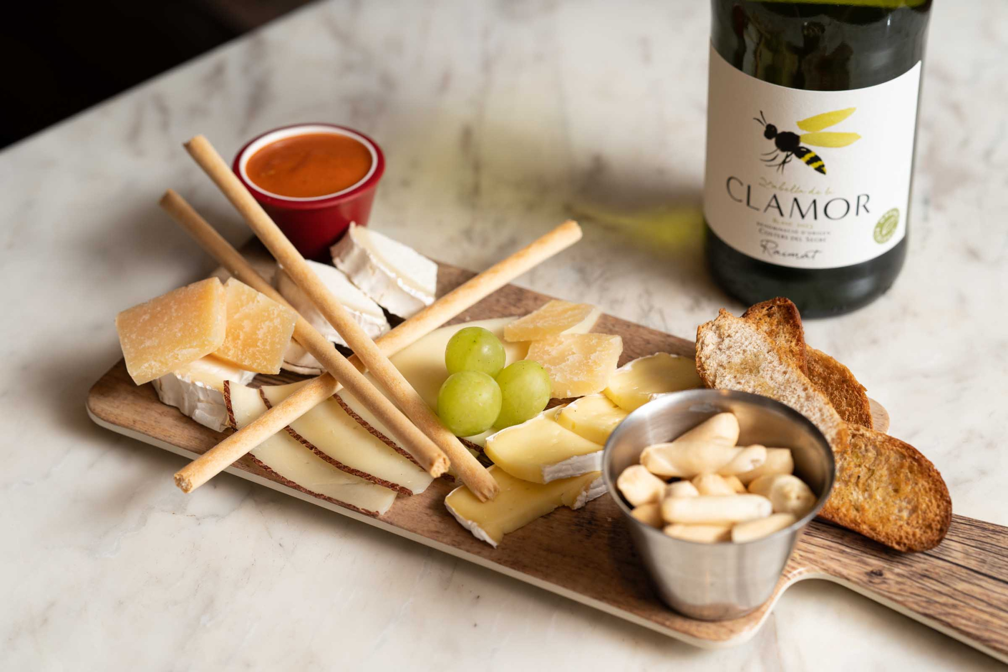 Se observan una tabla con aperitivos y brochetas, un cuenco con frutos secos y una botella de vino sobre una mesa.