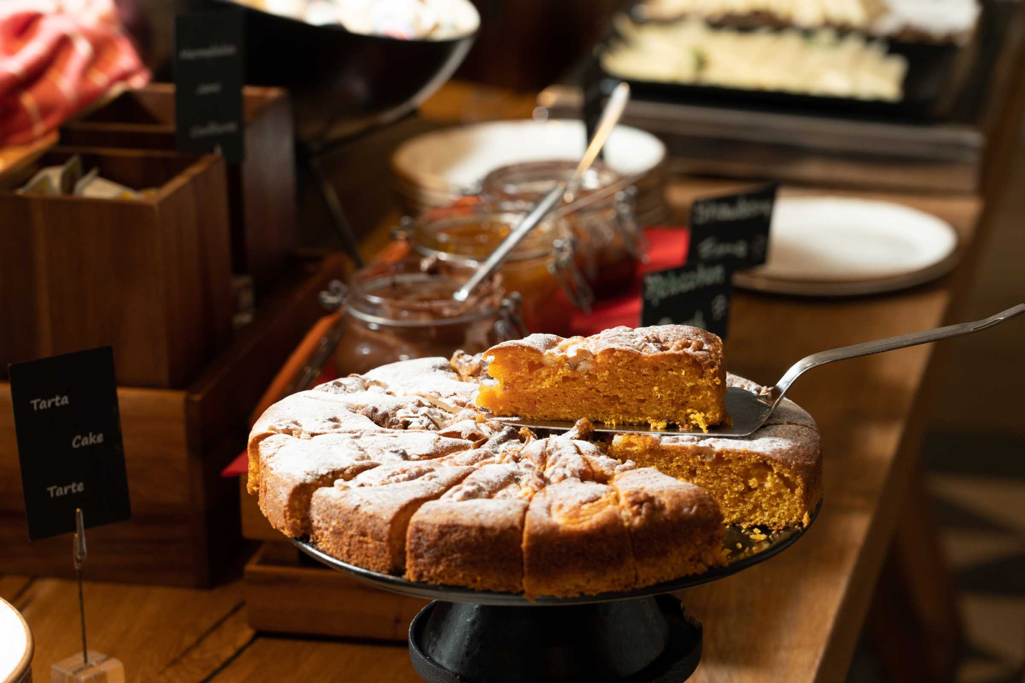 Se ve una mesa de buffet con pasteles y bandejas, con un letrero de opciones cerca.