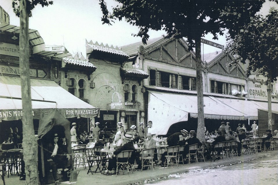 Se ven mesas y sillas al aire libre junto a una terraza cubierta en una calle con edificios al fondo.