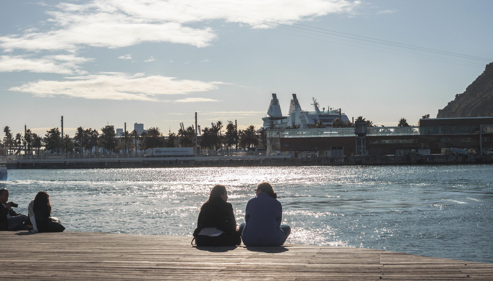 Dos personas se sientan en un paseo de madera junto al agua, con barcos y una grúa al fondo.
