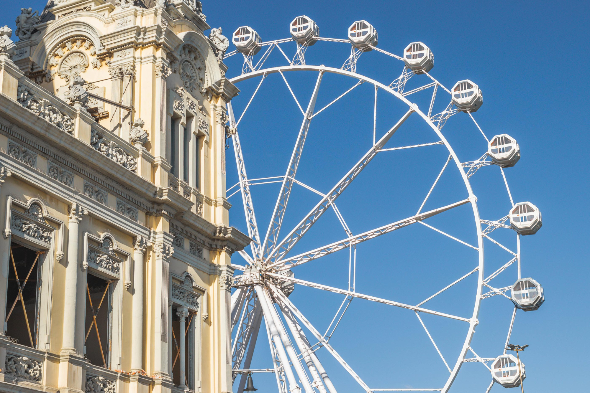 Muestra una rueda de la fortuna gigante sobre el cielo azul, junto a una fachada histórica de edificios.