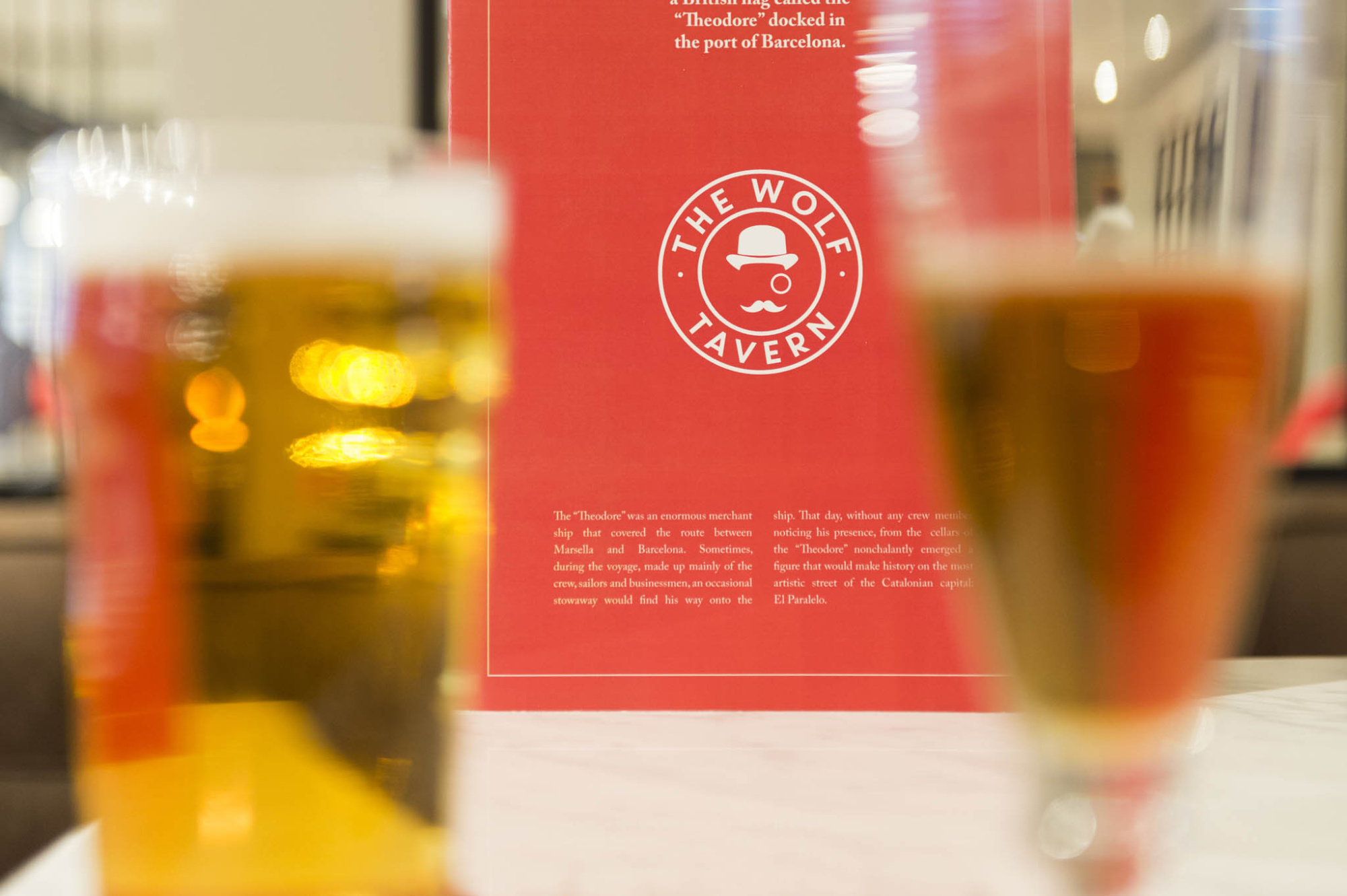 Vasos con bebidas y un cartel rojo con el texto “the wolf tavern” aparecen sobre una mesa en un bar.