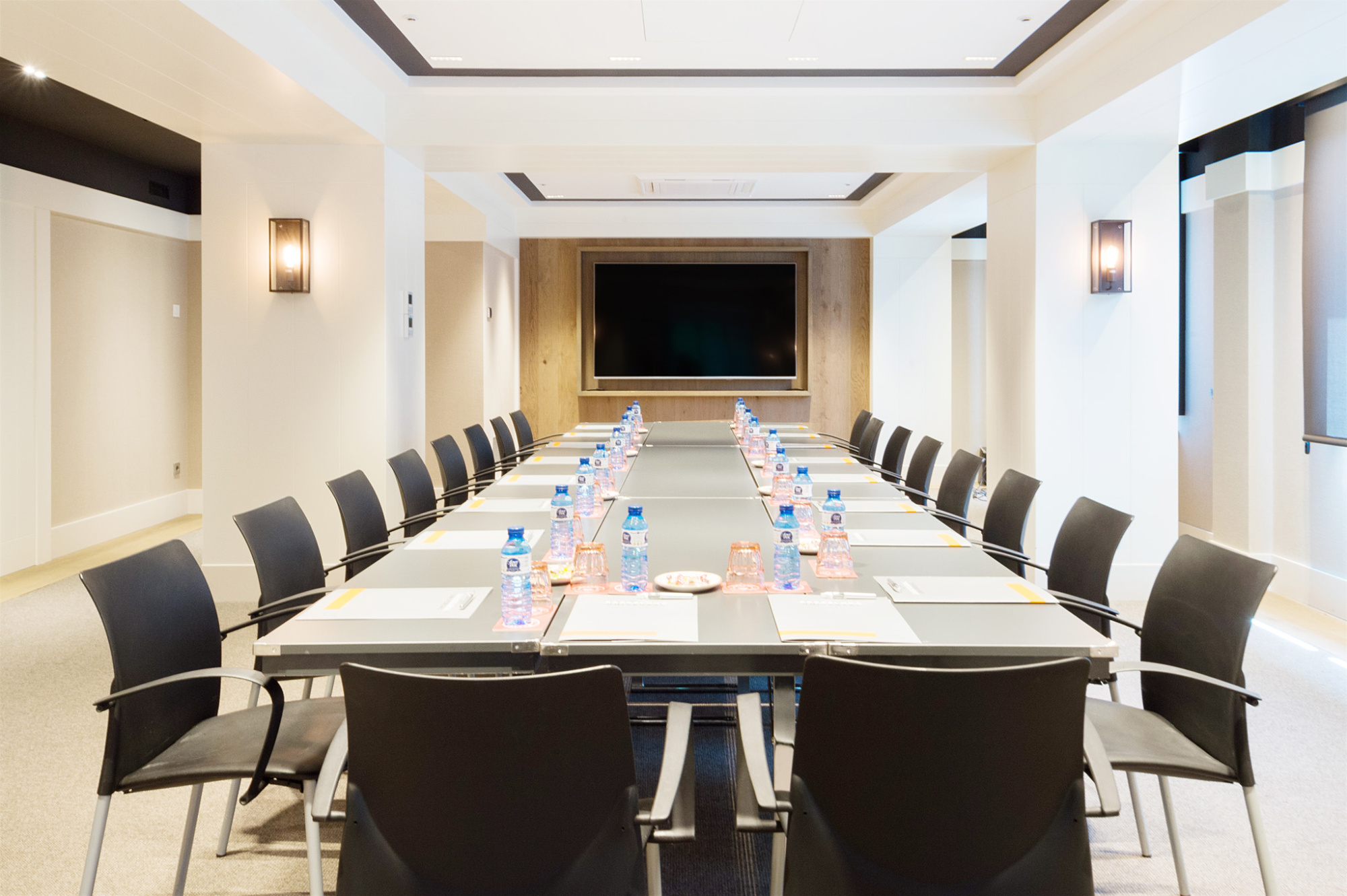 Se ve una sala con mesa de conferencias y sillas alrededor, con paredes blancas y luz empotrada.