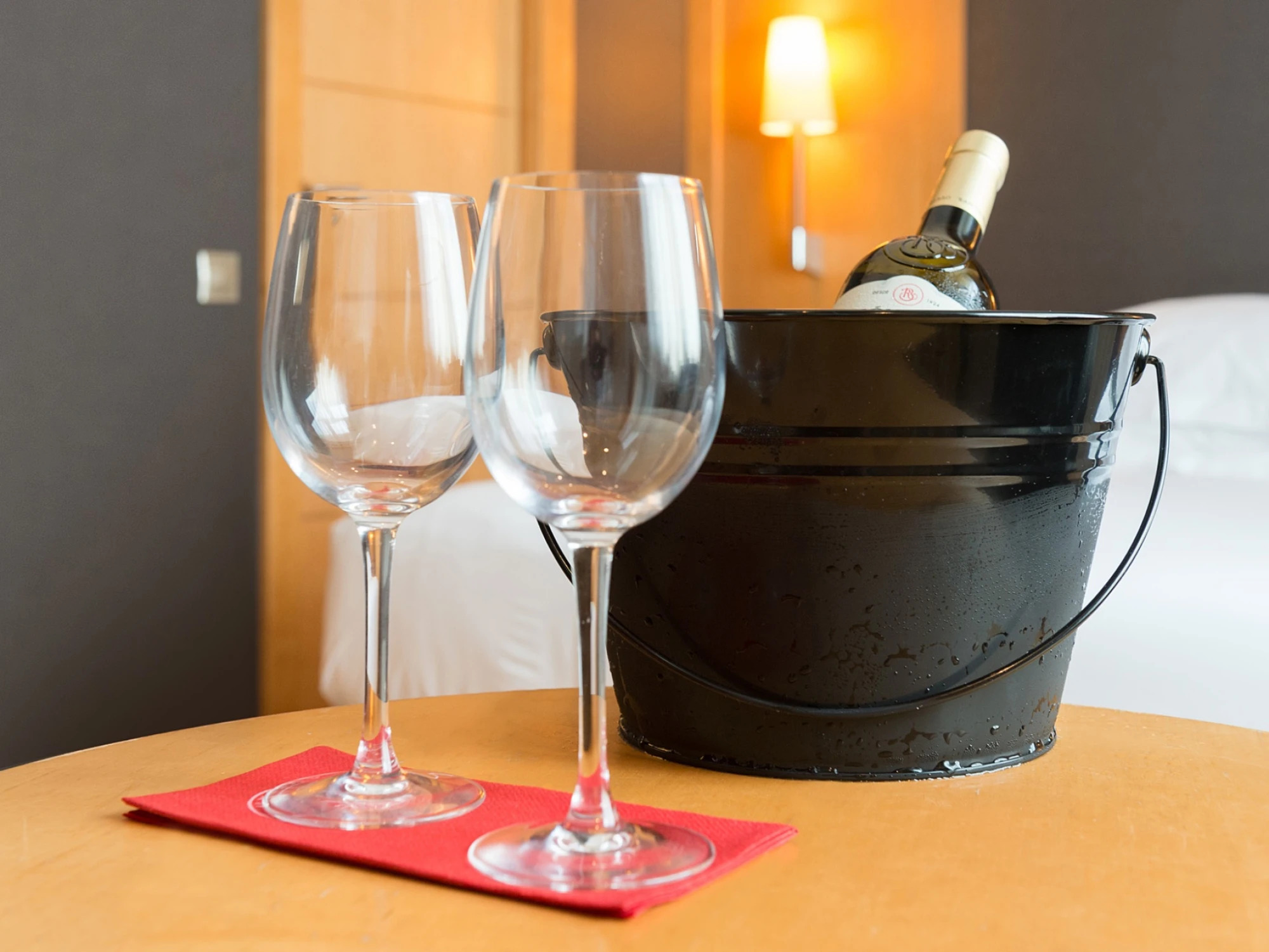Se ven dos copas de vino y una cubitera con una botella sobre una mesa junto a un entorno interior iluminado.